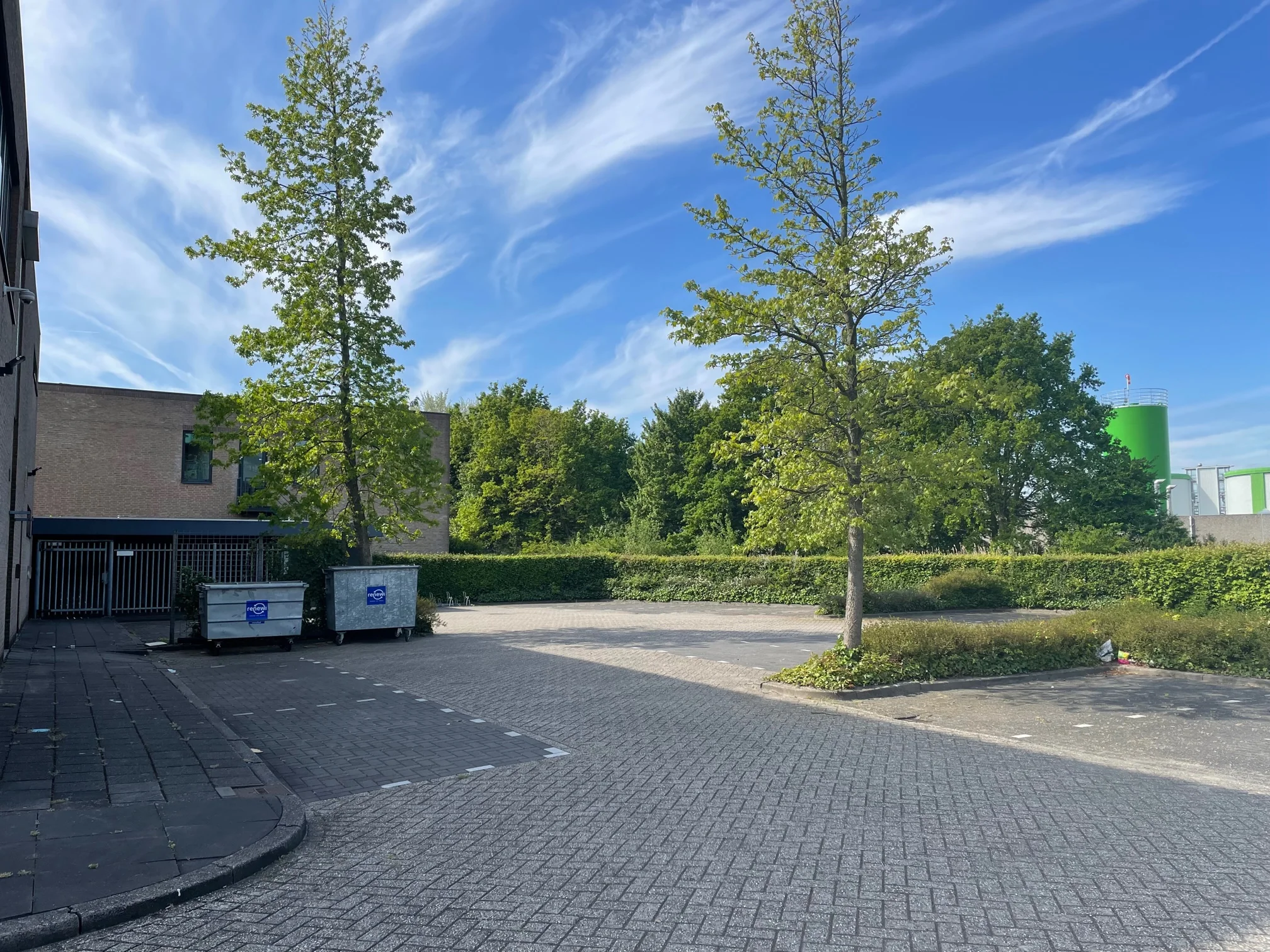 Leeg parkeerterrein aan de Rivium 4e Straat met bomen, afvalcontainers en een industrieel gebouw op de achtergrond.